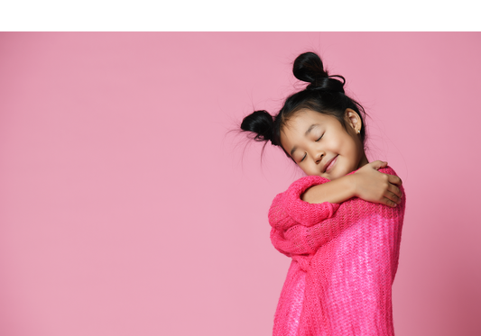 Young, dark-haired girl in pink sweater hugging herself in front of soft pink background.